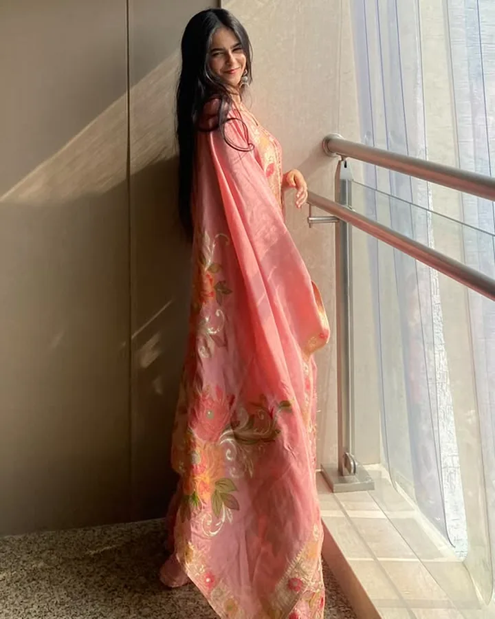 Himanshi in a coral saree, standing by a window